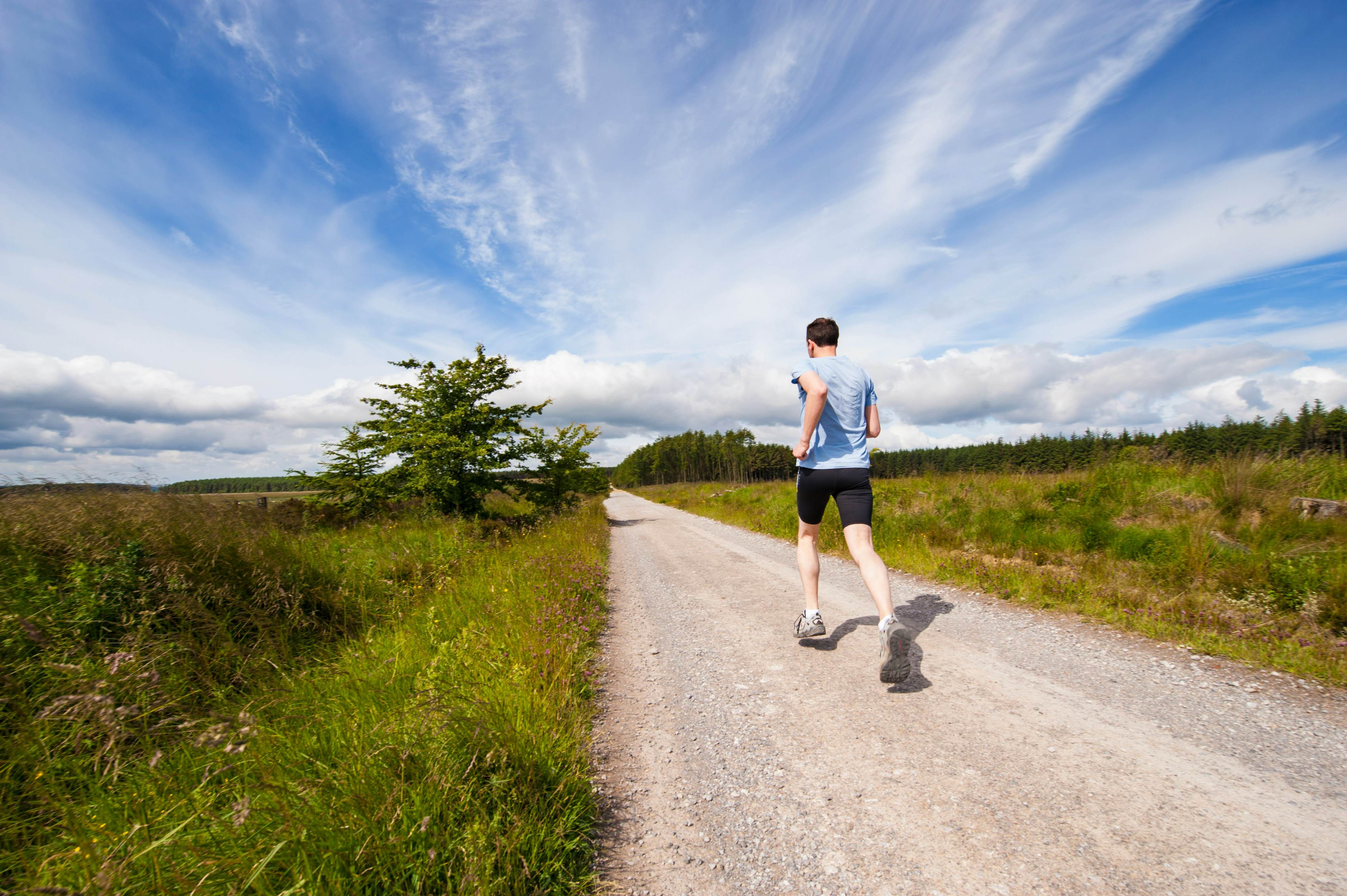 A man running on a gravel path surrounded by lush greenery and a bright blue sky, illustrating outdoor running and fitness improvement tips from Mojo Fitness Littleton.